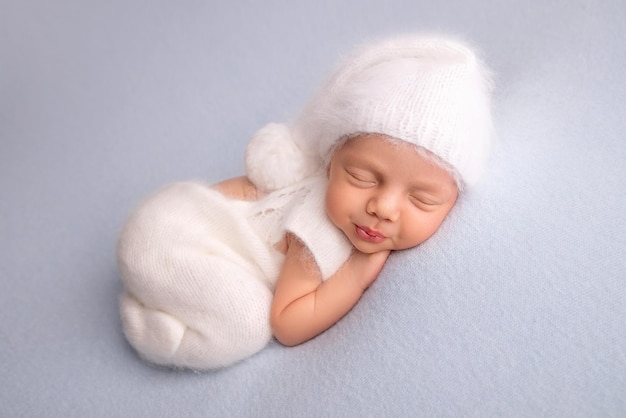 Sleeping newborn girl in the first days of life in a white soft bodysuit with a knitted woolen white cap on a blue background. Studio macro photography, portrait of a newborn. Woman's happiness.