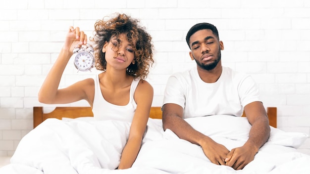 Sleepy africanamerican couple sitting in bed with clock