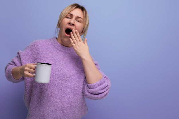 Sleepy blond young millennial woman in a casual look invigorates with a cup of coffee on a bright