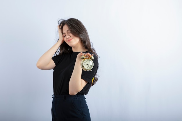 sleepy girl holding alarm clock on white wall. 