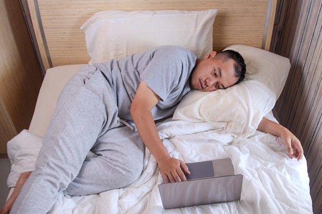 Sleepy lazy man working on laptop while sleeping on a bed