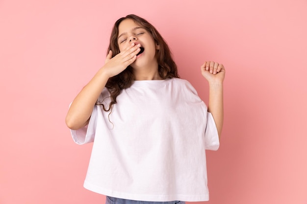 Sleepy little girl wearing white Tshirt standing and yawning with closed eyes and raised arms