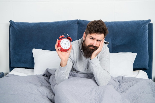 Sleepy man holding alarm clock being in bed in morning time