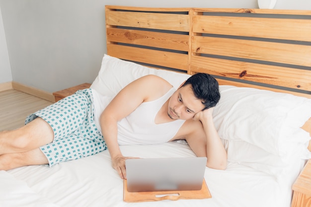 Sleepy man is working with his laptop on his cozy bed. Concept of boring freelancer lifestyle.