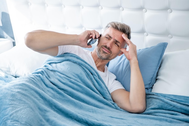 Sleepy mature man in bed speaking on phone headache