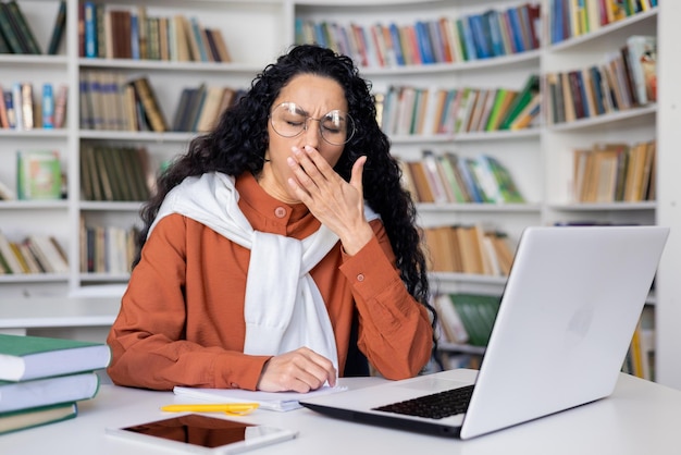 Sleepy and overtired young student studying in campus academic library until late hispanic woman