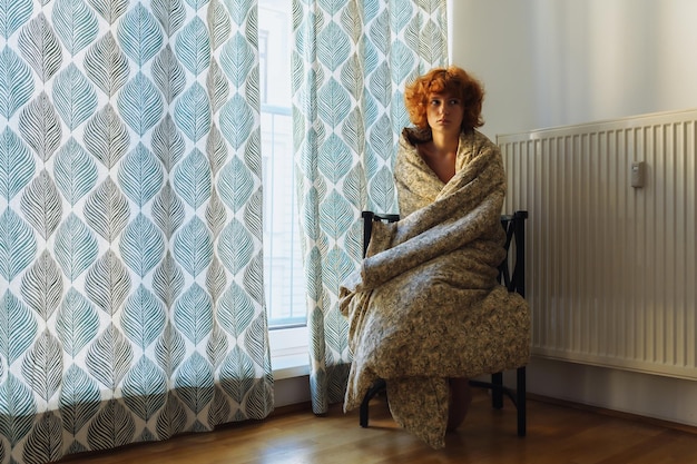 Sleepy teenage girl in blanket sitting on chair near radiator