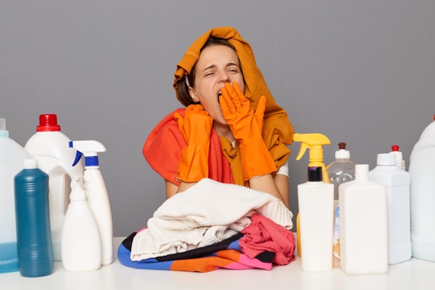 Sleepy tired woman posing at workplace with cleaning detergents isolated over gray background yawning covering mouth with hands being exhausted working long hours