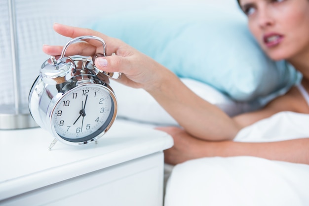 Sleepy young woman in bed extending hand to alarm clock 