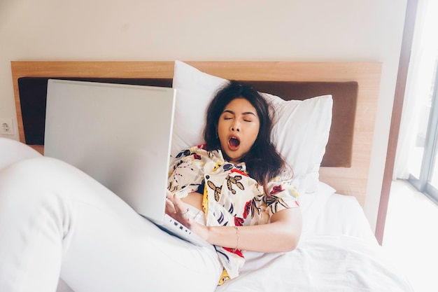 A sleepy young woman yawning while working from the bed