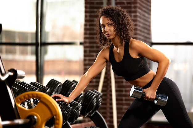 A slender athletic African American woman in sportswear does exercises with dumbbells in the gym against a mirror The concept of sports and recreation