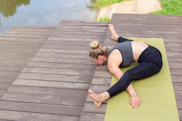 A slender blonde woman on a wooden platform on mat does yoga and stretching