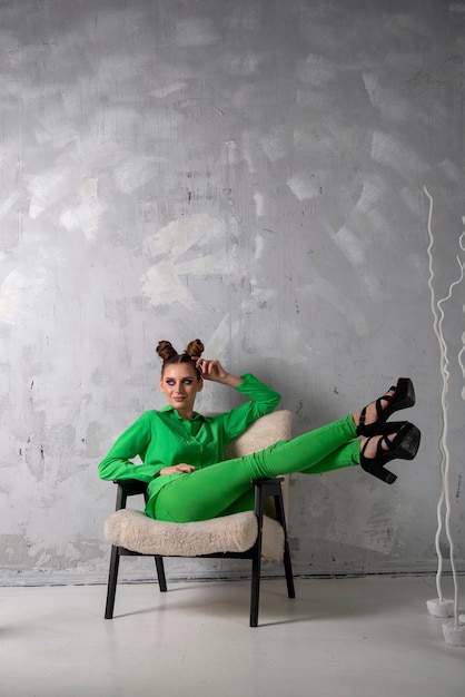 Slender girl on heels and in stylish green suit sits on chair on gray wall background in studio Vertical frame