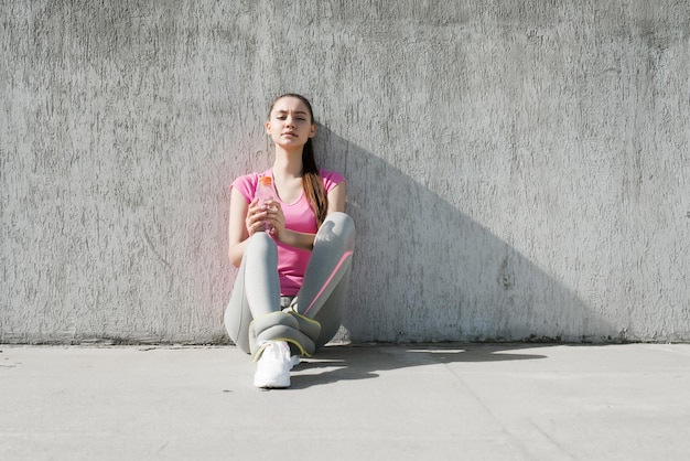 Slender tired young girl in a pink Tshirt sitting on the ground and resting after a workout