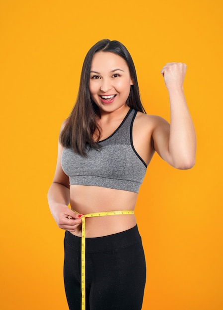 A slender woman measures her waist with a measuring tape. Against a yellow background.