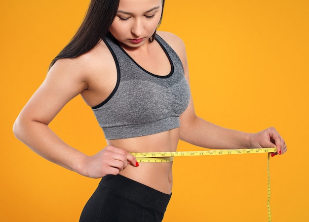 A slender woman measures her waist with a measuring tape. Against a yellow background.