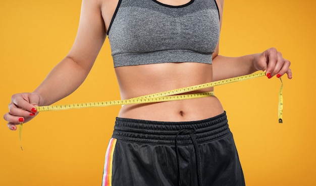 A slender woman measures her waist with a measuring tape on a yellow background.