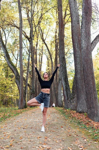 Slender woman performs vrikshasana exercise tree pose with arms raised and spread to the sides