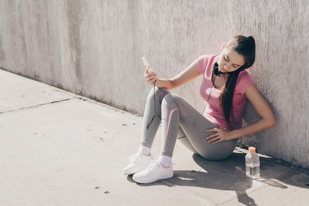 Slender young girl resting after a difficult workout outdoors sitting on the ground and listening to music on headphones