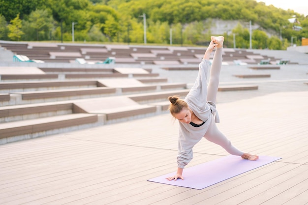 Slender young serene woman performing advanced asana of yoga outside standing on fitness mat outdoors