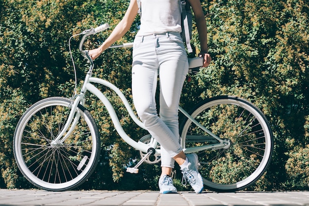 Slender young woman in a white T-shirt and jeans stands near her stylish bicycle in the summer