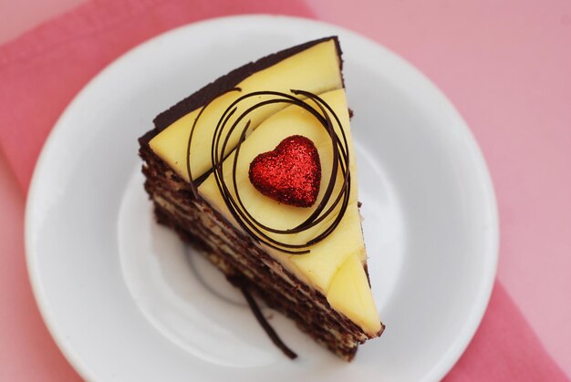 A slice of Delicious Chocolate Cake with Yellow Cream and Red Heart on a plate. Pink Background