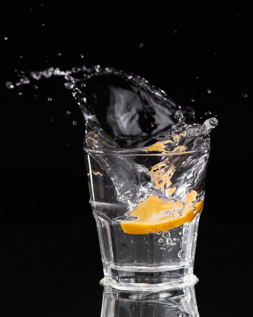 Slice of lemon splashing into a glass of water with a spray of water droplets in motion suspended in the air above the glass on a dark background.