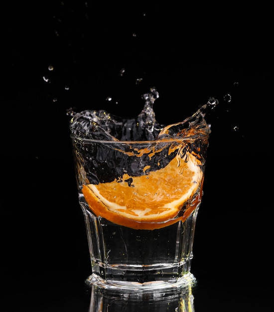 Slice of lemon splashing into a glass of water with a spray of water droplets in motion suspended in the air above the glass on a dark background.