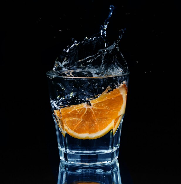 Slice of lemon splashing into a glass of water with a spray of water droplets in motion suspended in the air above the glass on a dark background.