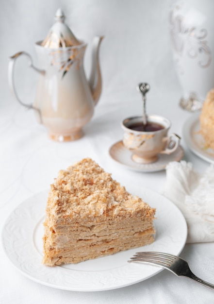 Slice of multilayer Napoleon cake with butter cream On a white plate Closeup Next to a plate is a napkin and a fork In the background is a cup teapot and a vase of flowers White background