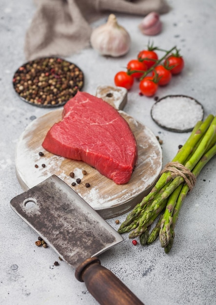 Slice of Raw Beef sirlion steak on round chopping board with tomatoes,garlic and asparagus tips and meat hatchet on light kitchen table background.