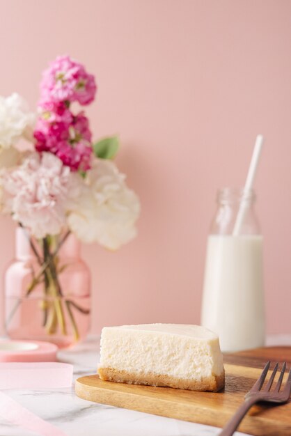 Slice of tasty homemade cheesecake with flowers and miilk on pink background. Healthy organic summer dessert pie side view vertical