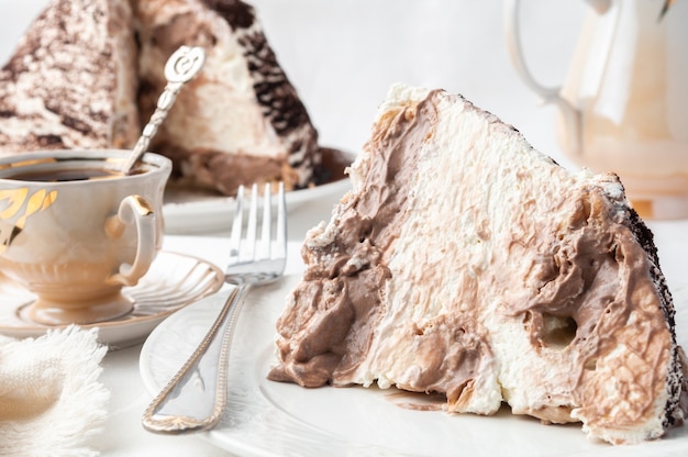 Slice of Tiramassu cake on a white plate Home made Near a fork and a cup of coffee In the background is a plate with cake and a coffee pot White background Closeup