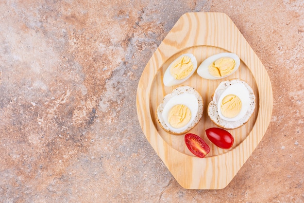 Sliced boiled egg, tomatoes and baguette bread on a wooden plate, on the marble background.