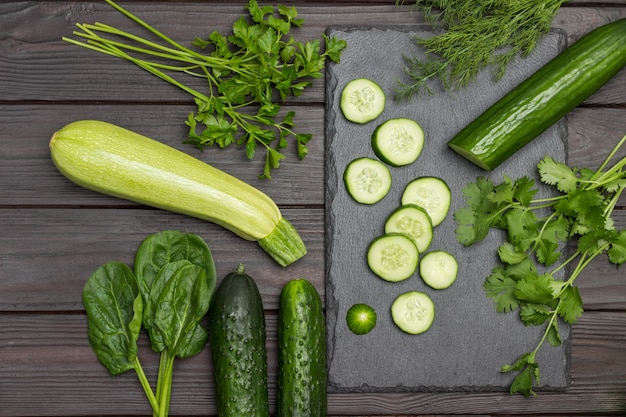 Sliced cucumber, cilantro and dill on cutting board. Zucchini, spinach and parsley on table. Dark wooden background. Flat lay.