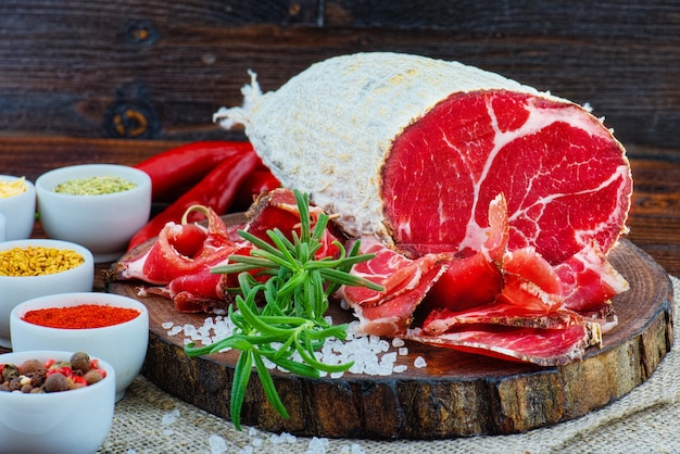 Sliced cured coppa with spices and a sprig of rosemary on dark wooden rustic background.