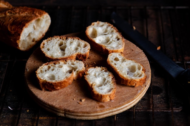 Sliced French baguette on a wooden board on a dark background Bread with big holes Preparing for bruschetta