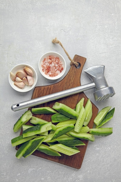 Sliced fresh cucumbers on a wooden cutting board kitchen hammer garlic and pink salt on a light concrete table