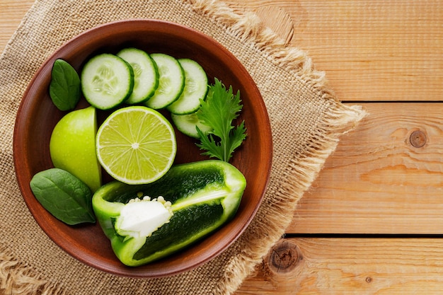 Sliced green fruits and vegetables on a wooden background. Chopped green apple, bell pepper, cucumber and lime in a clay bowl. Top view. Copy space