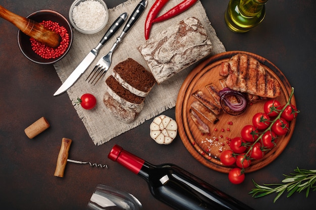 Sliced grilled pork steaks with bottle of wine, wine glass, corkscrew, knife, fork, black bread, cherry tomatoes, garlic