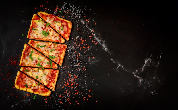 Sliced Hawaiian pizza on black stone, top view.