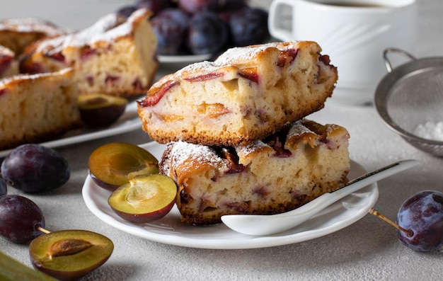 Sliced plum pie sprinkled with powdered sugar and a cup of coffee on a light gray table