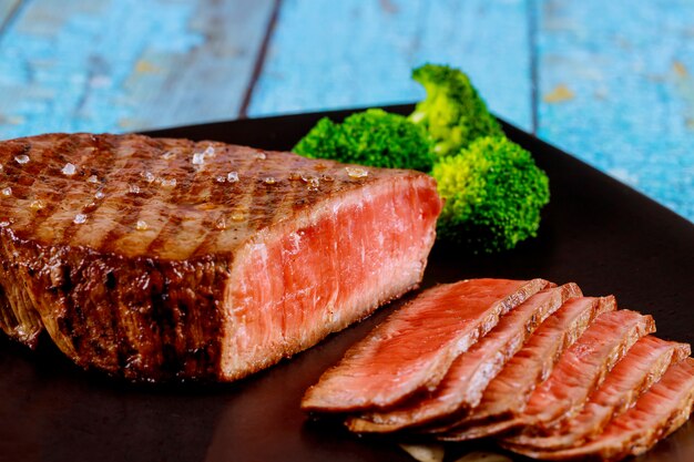 Sliced rare beef tenderloin with broccoli on black background.