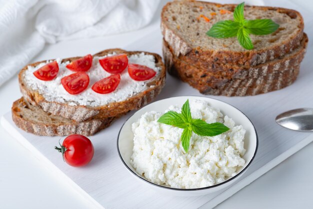 Slices of bread with cottage cheese and cherry tomatoes on wooden cutting board on white background