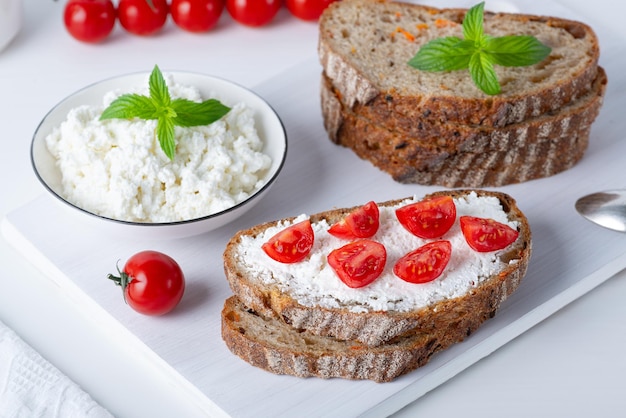 Slices of bread with cottage cheese and cherry tomatoes on wooden cutting board on white background