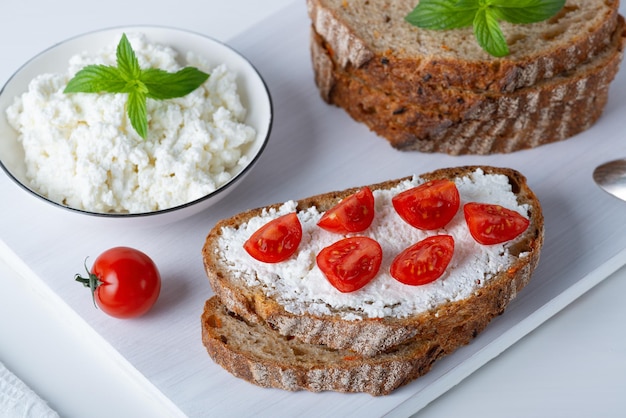 Slices of bread with cottage cheese and cherry tomatoes on wooden cutting board on white background