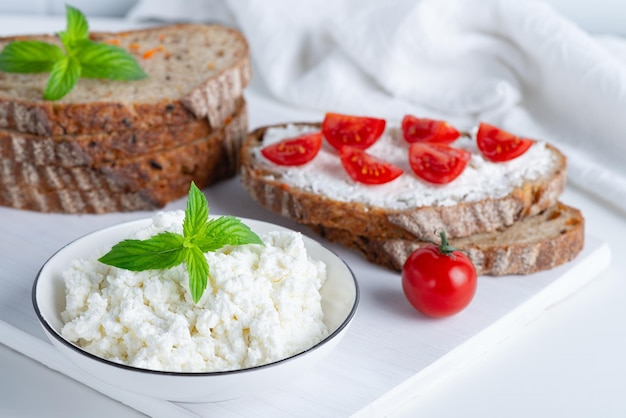 Slices of bread with cottage cheese and cherry tomatoes on wooden cutting board on white background