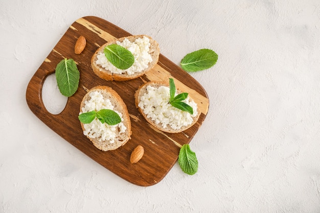 Slices of bread with cottage cheese and mint leaves on wooden cutting board on white background. Top view. Copy space.