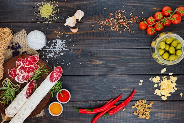 Slices of chopped sausage, spices in jars and scattered, pepper chili and tomatoes on a dark wooden rustic background.