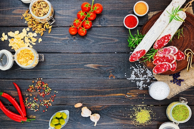 Slices of chopped sausage, spices in jars and scattered, pepper chili and tomatoes on a dark wooden rustic background.